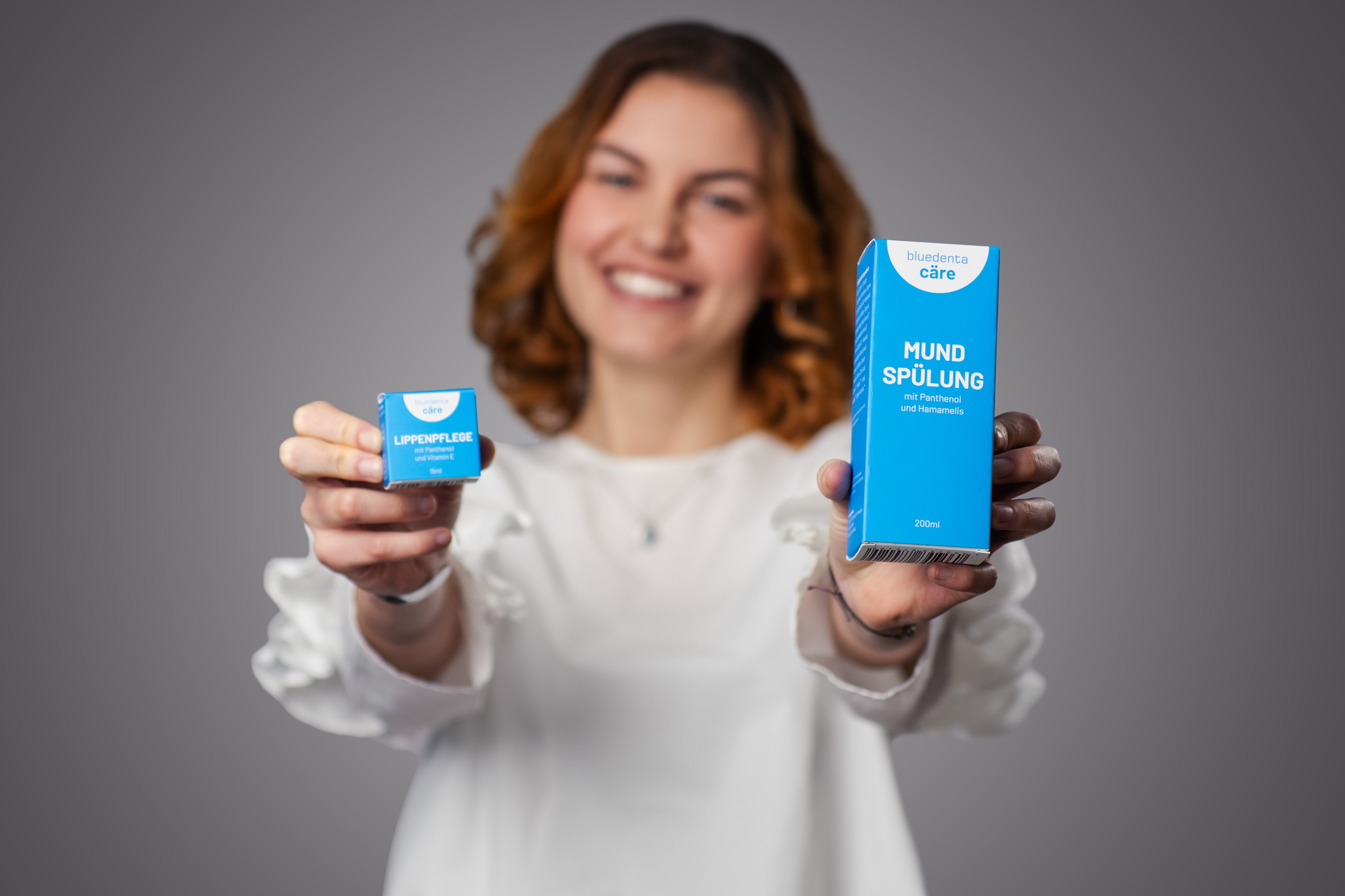 Eine junge rothaarige Frau in weisser Bluse hält prominent zwei Produkte der Marke bluedenta cäre in den Bildvordergrund. Es handelt sich um die himmelblauen Produktkartons der Mundspülung und Lippenpflege von bluedenta cäre.