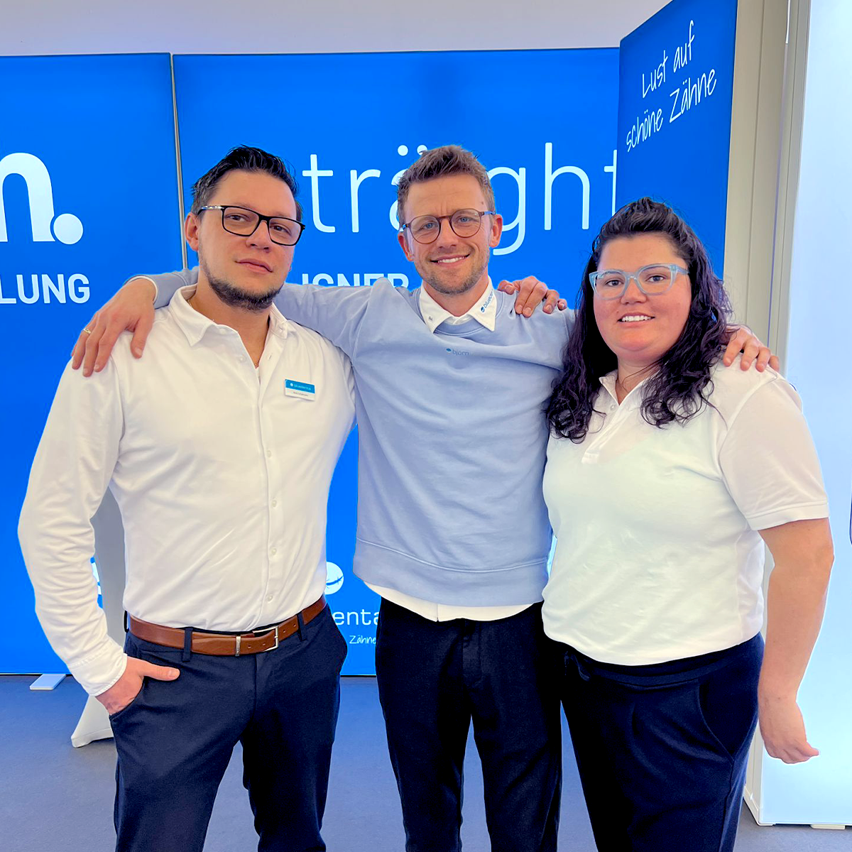 Das bluedenta Team, 2 Männer und eine Frau, stehen mit breitem, sympathischem Lächeln vor dem himmelblauen bluedenta Messestand.