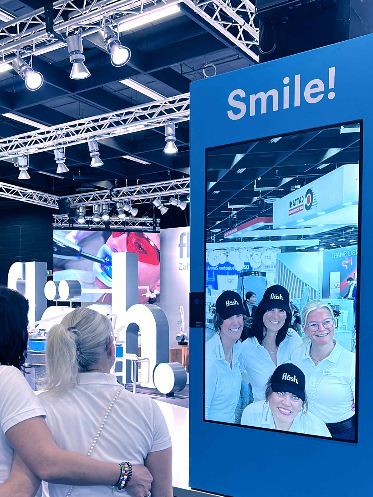 bluedenta auf der IDS 2025! 4 junge Dentalhygienikerinnen posieren vor der fläsh Smile Cam. Ihr von der fläsh App digital bearbeitetes Bild mit weissem Lächeln und fläsh Baseballkappe auf dreien der vier Köpfe ist in einem MEssescreen zu sehen.