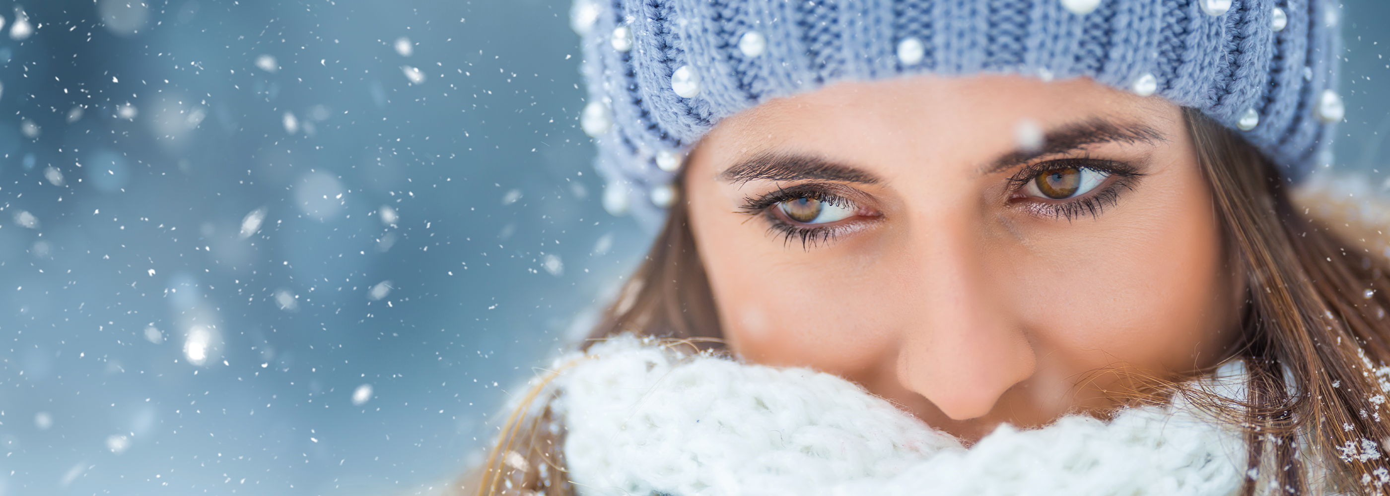 Nahaufnahme einer jungen Frau mit dunklen Augen, der Mund ist von einem weißen Wollschal verdeckt. Sie trägt eine hellblaue Wollmütze. Im Hintergrund tanzen Schneeflocken.
