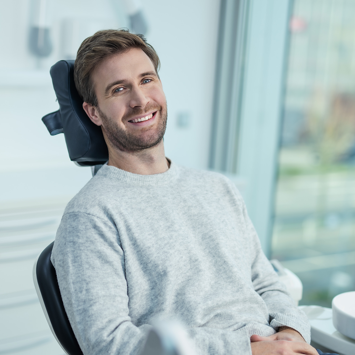 Ein junger Mann mir Dreitagebart sitzt lächelnd im Behandlungsstuhl einer Zahnarztpraxis.