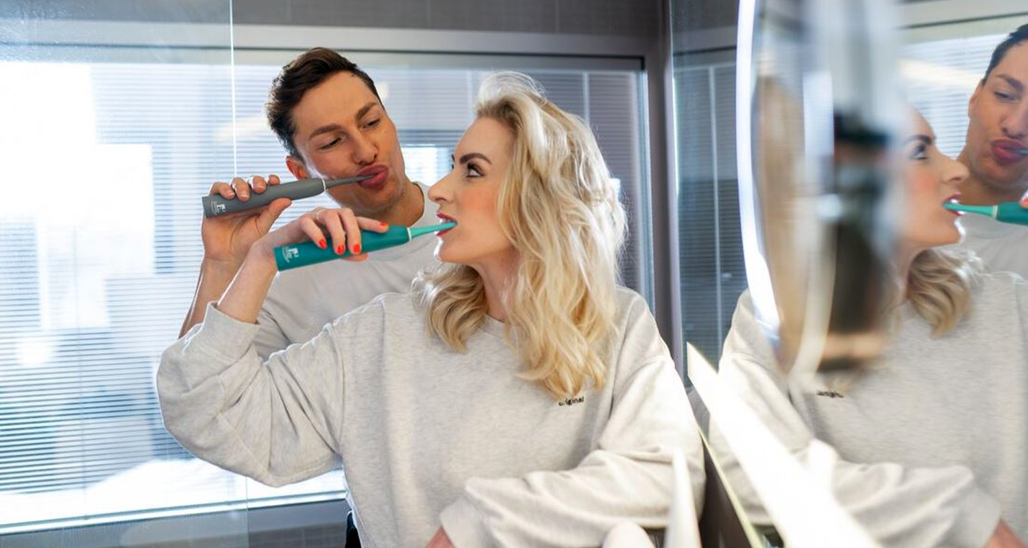 Ein junger, dunkelhaariger Mann und eine junge, blonde Frau in lässiger Casual Wear stehen gemeinsam an einem Badspiegel, der die rechte Seite des Bildes einnimmt. Beide putzen sich die Zähne mit einer sträight Schallzahnbürste. Er nutzt ein graues, sie ein grünes Modell. Sie steht vor ihm. Beide schauen sich beim Zähneputzen an.