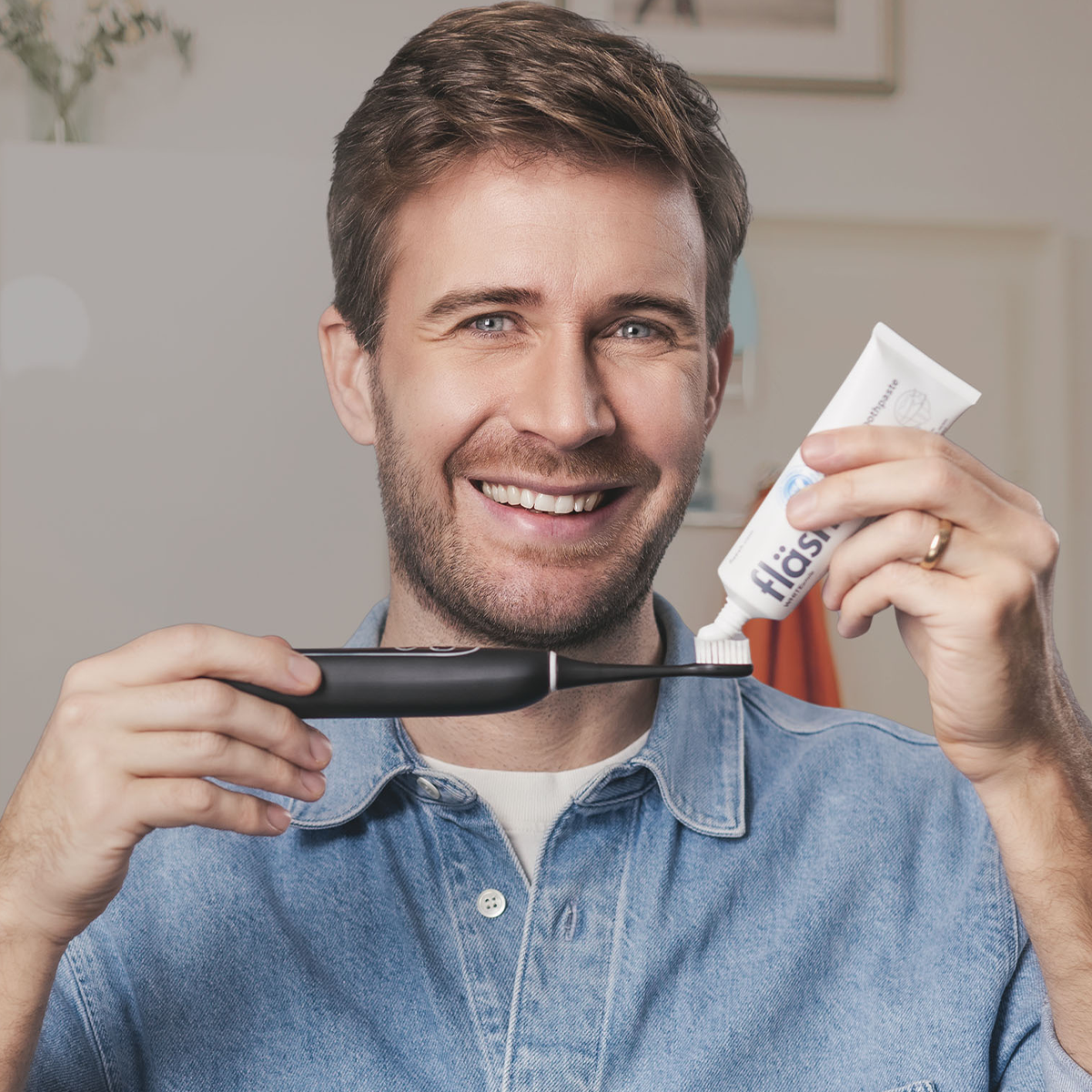 Ein junger Mann in Jeanshemd und mit sympathischem Lächeln gibt mit der linken Hand aus einer Tube fläsh Zahnpasta auf die sträight Schallzahnbürste, die er in der rechten Hand bereit hält.