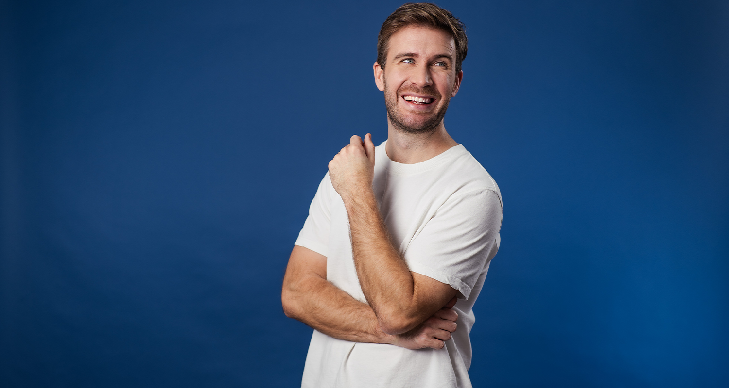 Eine junger Mann steht lässig in weissem Tshirt vor dunkelblauem Hintergrund. Er schaut nicht direkt in die Kamera, sondern zu einer nicht sichtbaren Person am rechten Bildrand und lacht dieser zu.