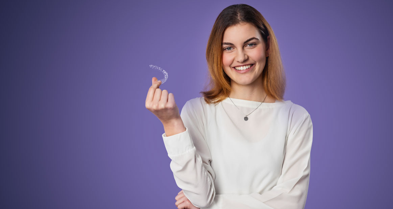 Eine junge, rothaarige Frau steht lächelnd vor lila Hintergrund. Sie hält in der rechten Hand eine transparente sträight Alignerschiene und lächelt dem Betrachter zu.