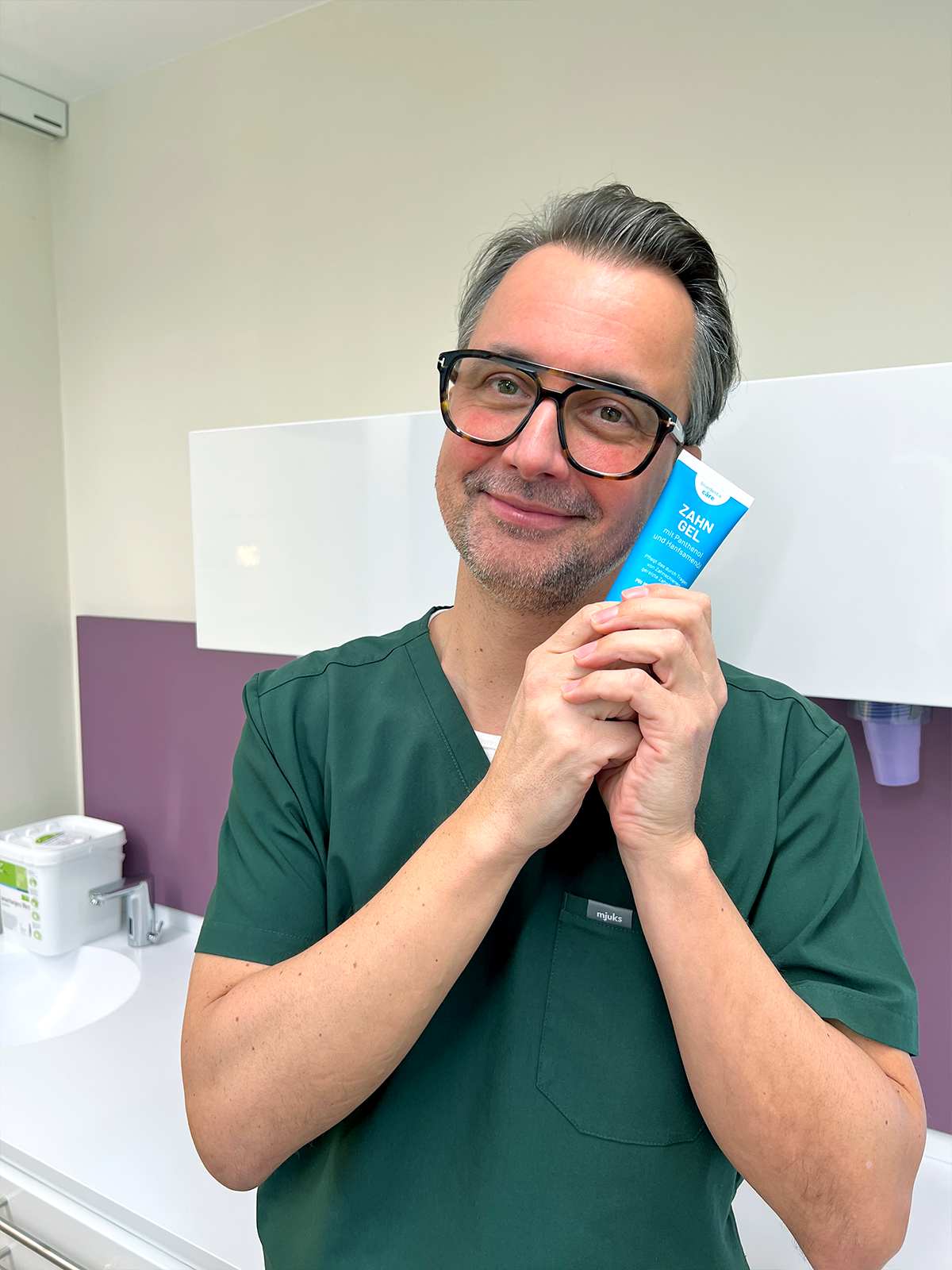 Zahnarzt Dr. Stephan Otto Lipphaus, steht in einem Behandlungszimmer seiner Praxis und hält liebevoll eine himmelblaue Tube bluedenta cäre Zahngel in die Kamera.
