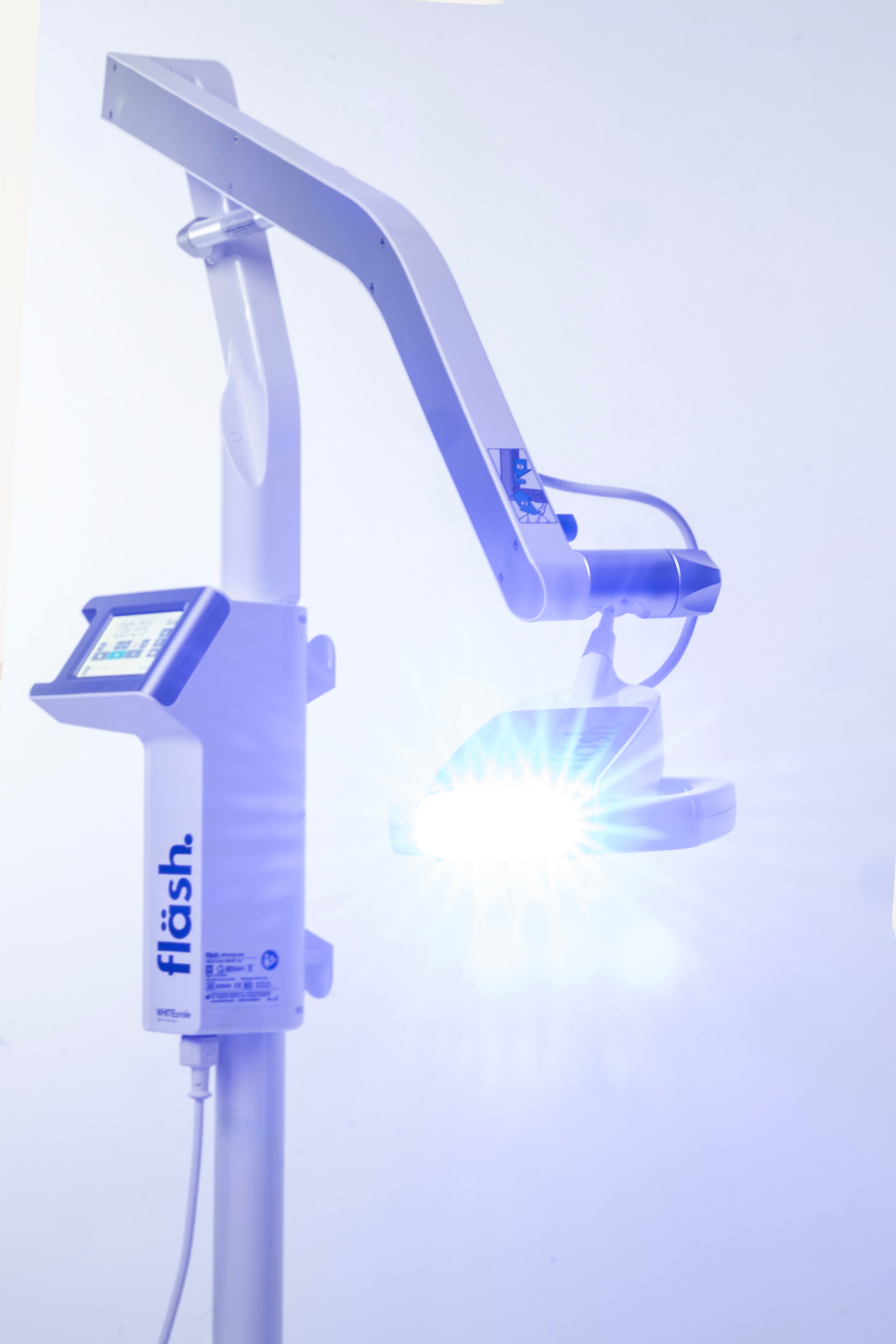 Vor weissem Hintergrund steht die fläsh Whitening Lampe, die für das professionelle Bleaching in Zahnarztpraxen verwendet wird. Die LED-Lampe leuchtet blau.