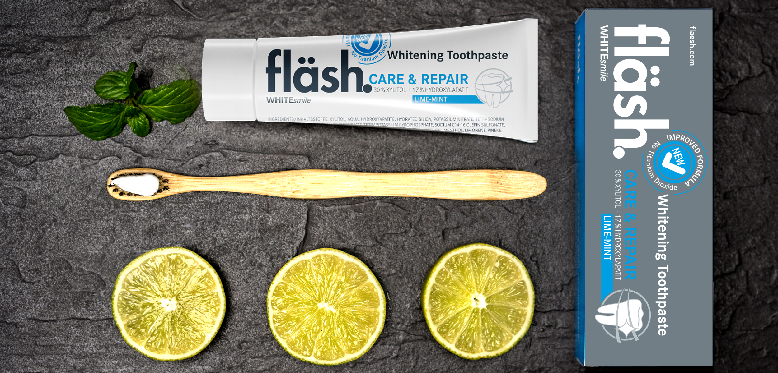 Auf einer grauen Schieferplatte liegt eine Tube fläsh Zahnpasta der Sorte Limette-Minze, der dazugehörige graue Produktkarton, eine Handzahnbürste aus Bambus, darunter drei Scheiben aufgeschnittene Limette. Neben der fläsh Tube liegen grüne Minzblätter.