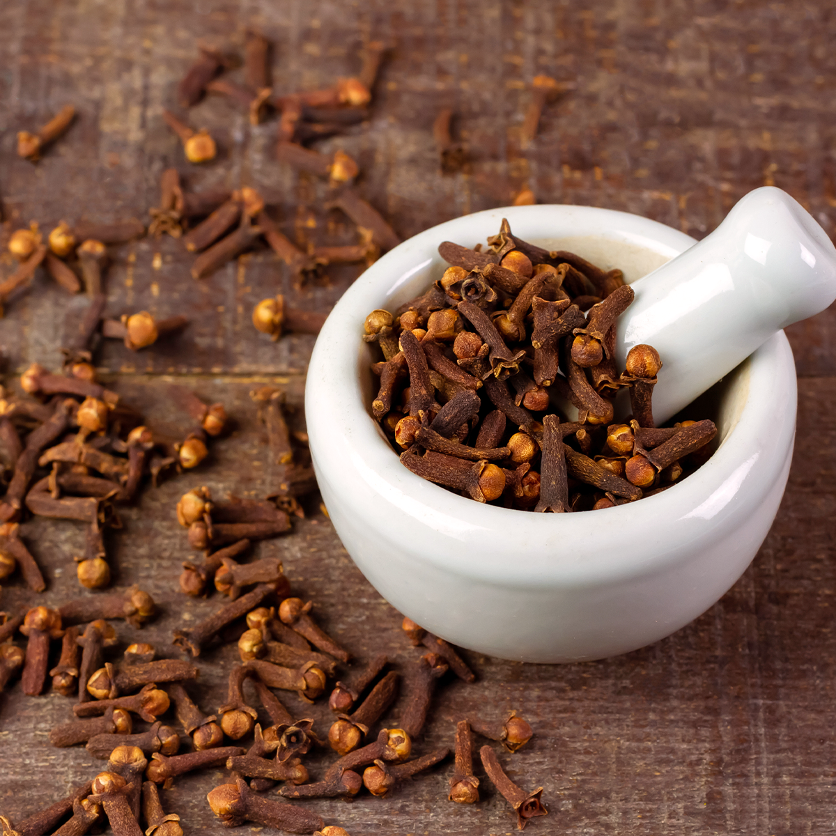 Eine Mörserschale aus weißem Porzellan mit dazu passendem Stößel steht auf einer Holzplatte. Im Mörser uns auf dem Holz liegen Gewürznelken.