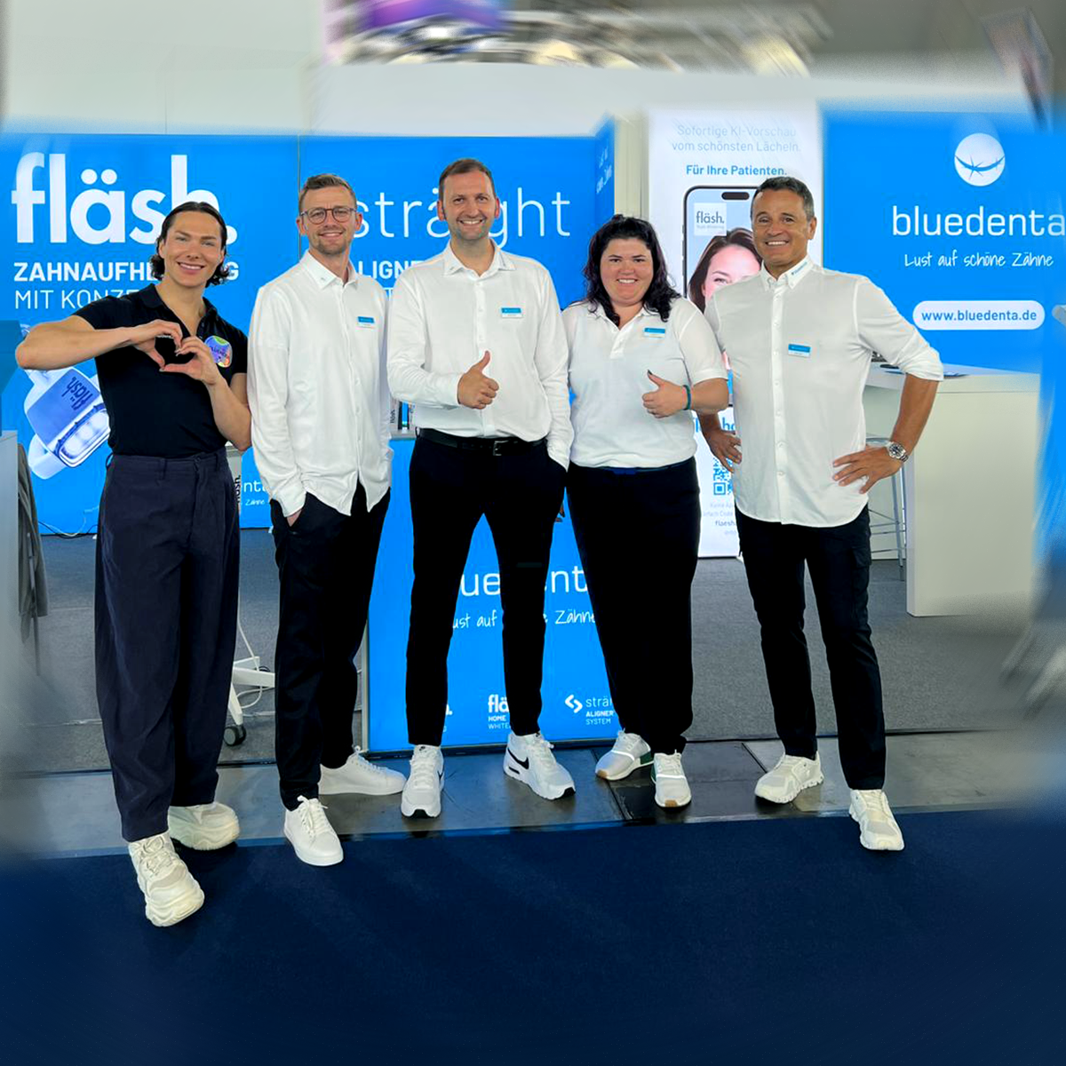 Das bluedenta Team, 5 Männer und eine Frau, stehen mit "Daumen hoch" oder "Herz"-Geste und breitem, sympathischem Lächeln vor dem himmelblauen bluedenta Messestand