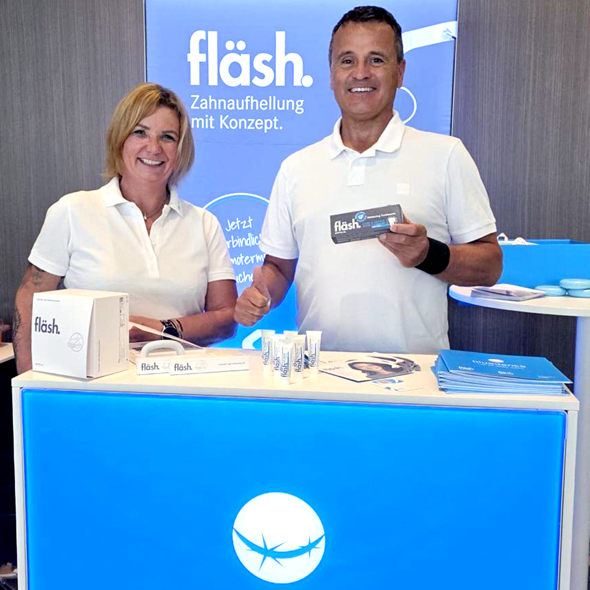 Das bluedenta Team, ein Mann und eine Frau, stehen mit sympathischem Lächeln vor dem himmelblauen bluedenta Messestand und präsentieren die Produkte von fläsh.