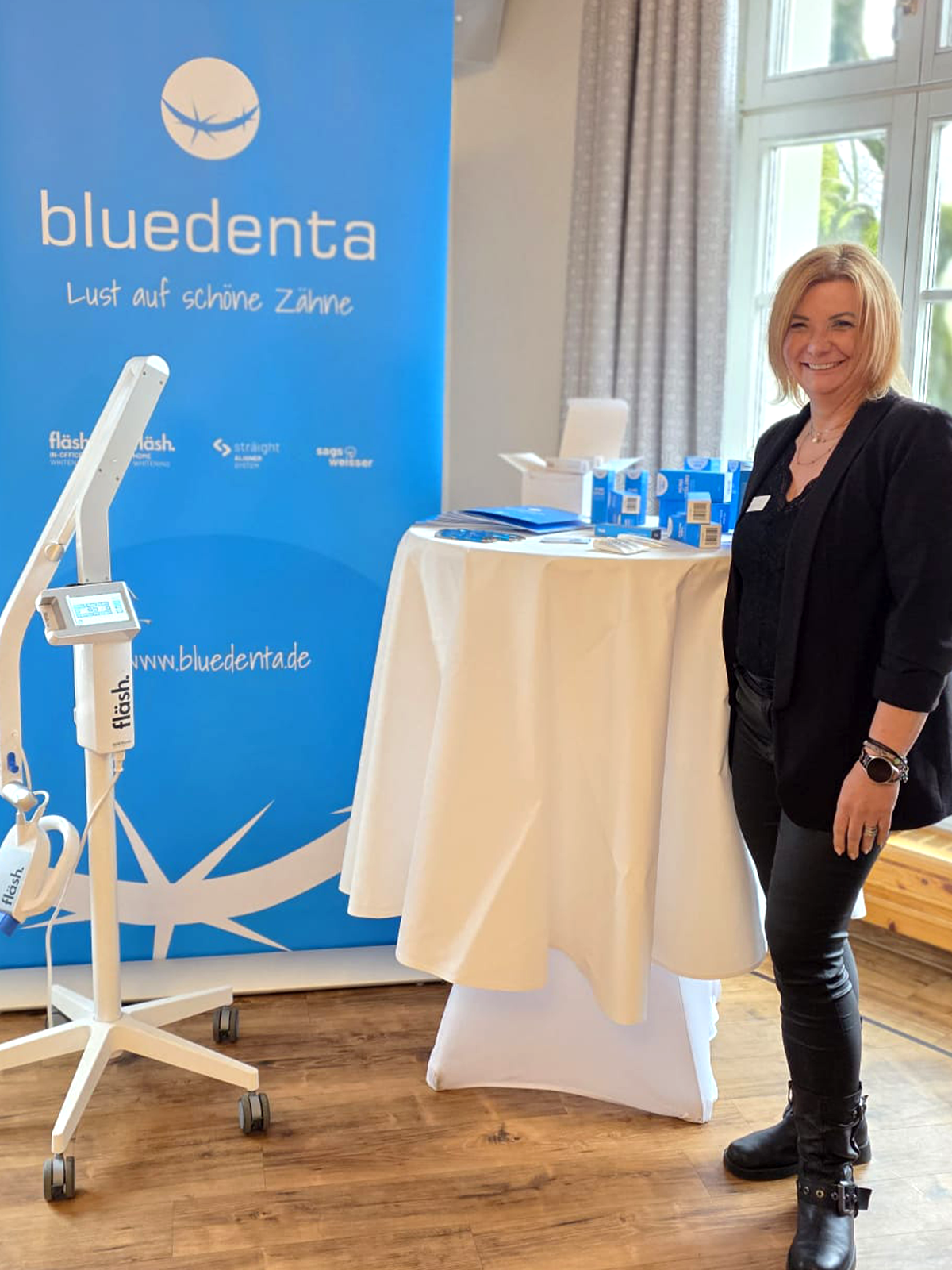 bluedenta Sales Managerin Katrin Keuenhoff steht lächelnd vor der himmelblauen bluedenta-Messewand mit einer fläsh Whitening Lampe. Auf einem Tisch stehen die Pflegeprodukte von bluedenta cäre. 