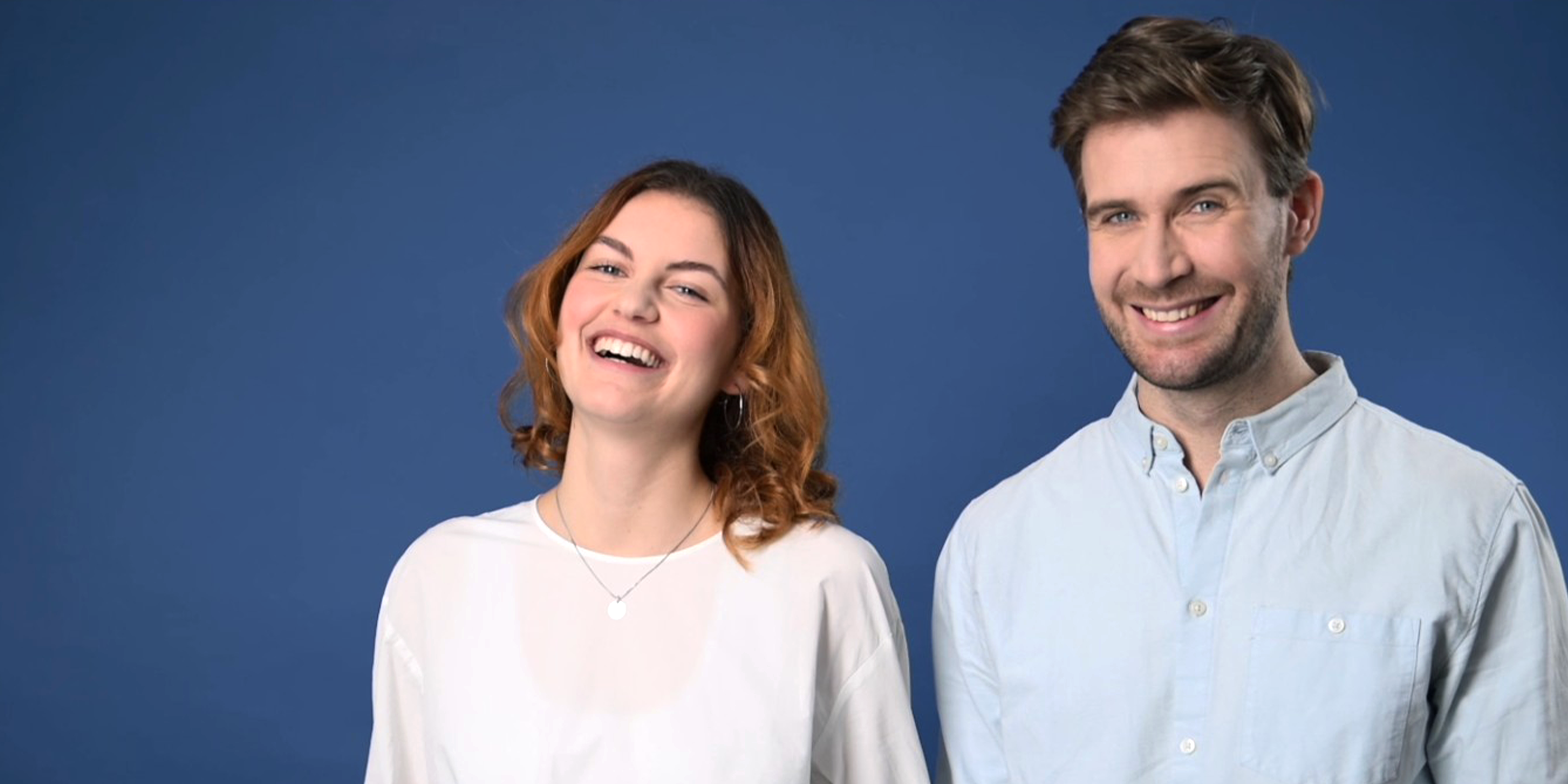 Eine junge, rothaarige Frau in weisser Bluse und ein junger Mann in hellblauem Hemd stehen nebeneinander vor dunkelblauem Hintergrund. Beide lachen ansteckend sympathisch in die Kamera.