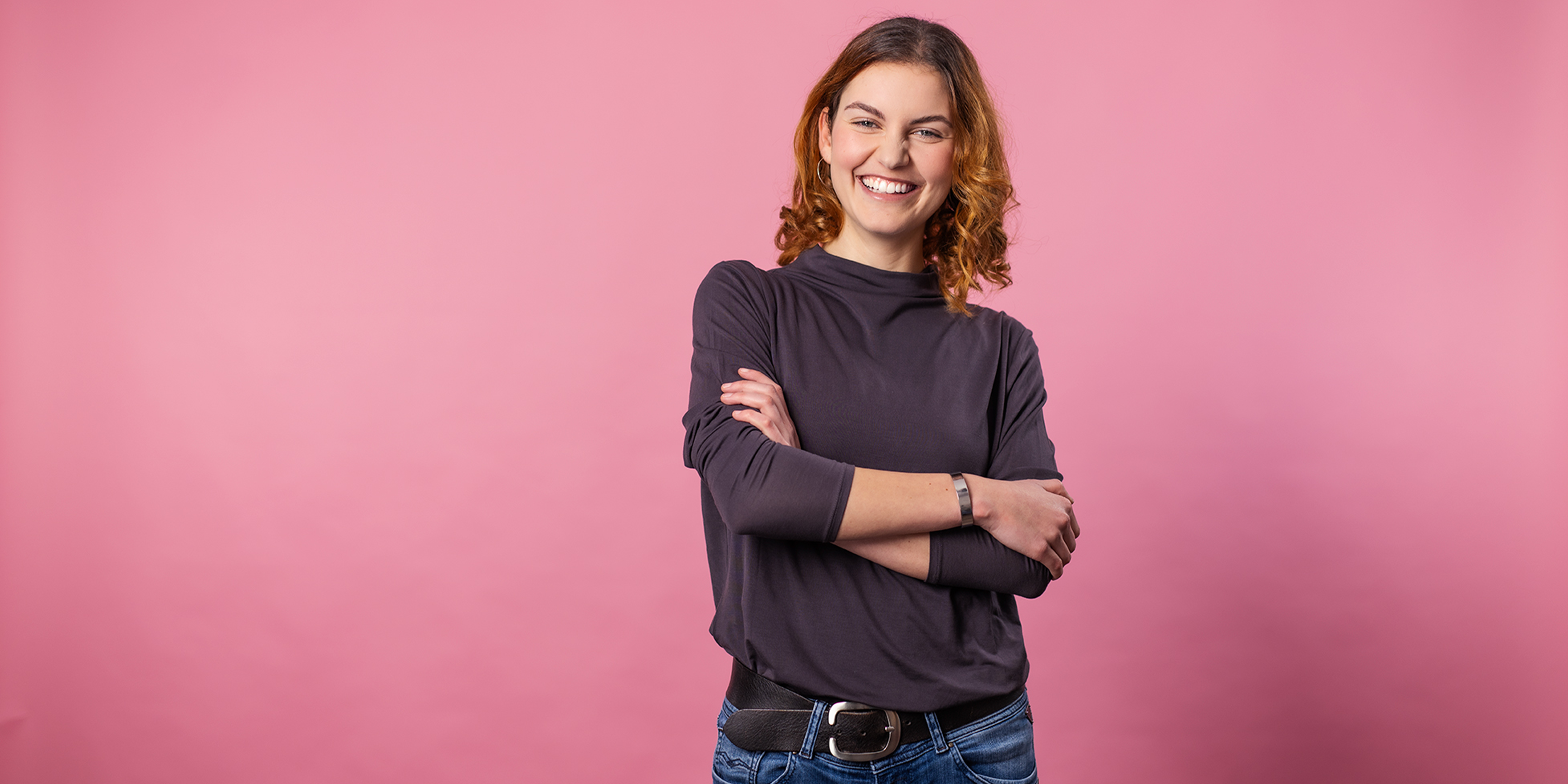 Eine junge rothaarige Frau in dunkelgrauem Shirt und blue Jeans steht vor altrosa Hintergrund. Sie wirkt selbstbewusst durch ihre vor dem Bauch verschränkten Arme und ihr offenes, sympathisches Lachen. Sie schaut direkt in die Kamera .