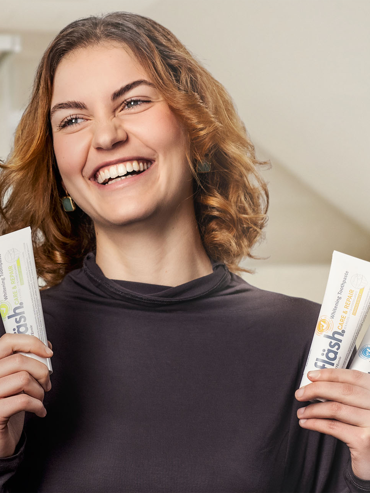 Eine rothaarige junge Frau lacht ansteckend sympathisch in die Kamera. Sie hält in der rechten Hand eine Tube fläsh Zahnpasta Kokos, in der linken eine Tube fläsh Zahnpasta Orange und eine Tube der Sorte Limette.