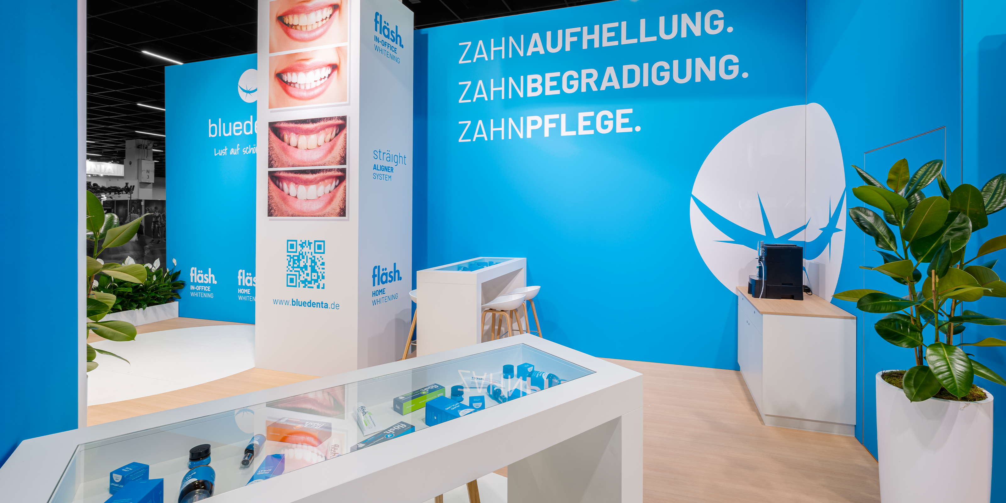 Blick auf den bluedenta Messestand auf der FIBO 2025. Auf himmelblauen Wänden steht in weissen Großbuchstaben die Essenz von bluedenta: "Zahnaufhellung. Zahnbegradigung. Zahnpflege." Auf einer weissen Säule sind Bilder von Zähnen vor und nach einem Bleaching mit fläsh zu sehen. Im Vordergrund ist ein gläserner Tresen, in dem die Zahnpflegeprodukte von bluedenta cäre präsentiert sind.